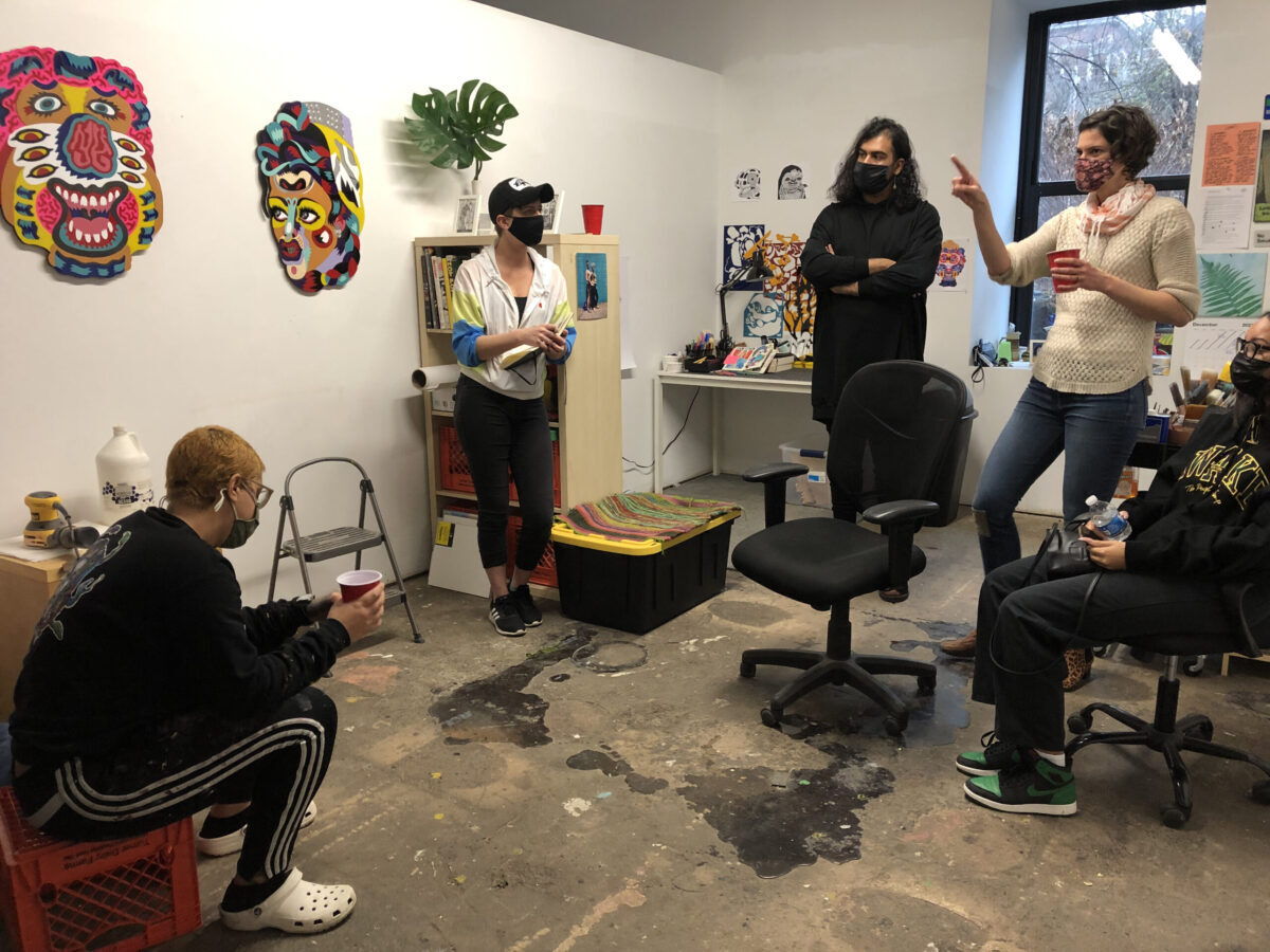 Four artists casually hang in a studio space with masks. One is pointing at artwork on the wall.