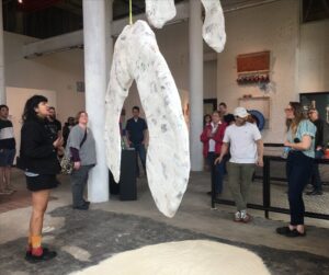 Artist stands next to their hanging sculpture of a giant seed pod.